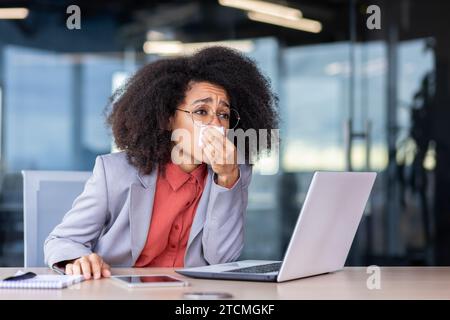 Afroamerikanerin, die im Büro arbeitet, hat eine laufende Nase, bläst ihre Nase in ein Taschentuch, sitzt an einem Tisch vor einem Laptop. Stockfoto