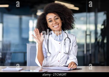 Afroamerikanische Ärztin berät Patienten per Videoanruf, sitzt an einem Tisch in einem privaten Büro, winkt in Begrüßung, Webcam-Ansicht. Stockfoto