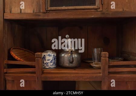Innenraum der vietnamesischen Küche mit Porzellan-Teekanne, Aluminiumkessel, Glasbecher, blauer Porzellanplatte, Rattan-Untersetzer in einem alten Holzschrank Stockfoto