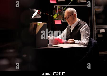 Ein älterer Privatdetektiv sitzt am Schreibtisch in einem dunklen Zimmer und arbeitet Überstunden in einem Strafverfahren. Leitender Polizist analysiert Vermisstenbericht, überprüft Beweisdokumente Stockfoto