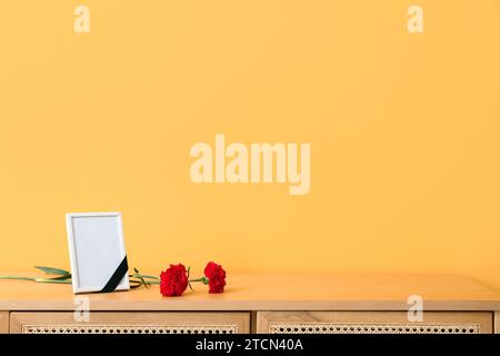 Leerer Trauerrahmen und Nelkenblumen auf Holzschrank in der Nähe der Farbwand Stockfoto