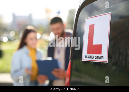 Fahrer und Kursleiter mit Klemmbrett in der Nähe des Autos im Freien, selektiver Fokus auf L-Platte. Fahrschule Stockfoto
