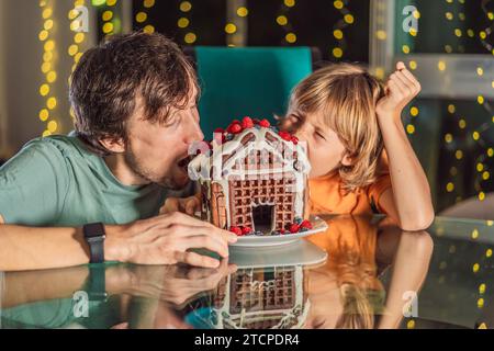 Erleben Sie einzigartige Momente, während Vater und Sohn in ein unkonventionelles Lebkuchenhaus beißen und die Weihnachtstraditionen eine neue Note verleihen. Eine leckere Mischung aus Kreativität Stockfoto