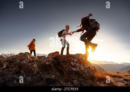 Gruppe junger Wanderer mit Rucksäcken geht mit Rucksäcken spazieren und hilft sich gegenseitig beim Klettern in den Bergen bei Sonnenuntergang Stockfoto