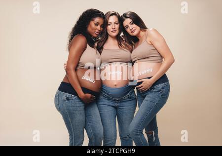 Junge, vielfältige schwangere Frauen im dritten Trimenon stehen zusammen in einem Studio. Sie tragen Jeans und BHs und zeigen ihre Babybeulen. Umarmt und los Stockfoto