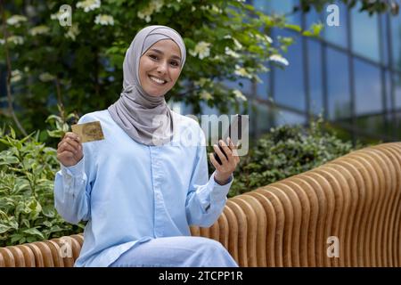 Glückliche attraktive Frau im Hijab, die draußen auf der Bank sitzt, Karte und Telefon hält, glücklich mit dem Einkaufen, lächelt und in die Kamera schaut. Stockfoto