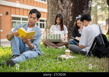 Ein fokussierter junger asiatischer Student liest ein Buch, während er mit seinen Freunden auf Gras sitzt. Bildungskonzept Stockfoto
