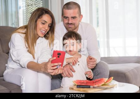 3-köpfige Familie mit einem kleinen Jungen, der das Handy benutzt, während er im Wohnzimmer Obst isst Stockfoto