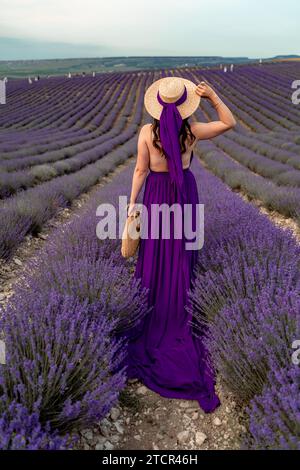 Ein junges, hübsches Mädchen in einem lila fliegenden Kleid steht auf einem blühenden Lavendelfeld. Rückansicht. Das Modell hat einen Strohhut Stockfoto