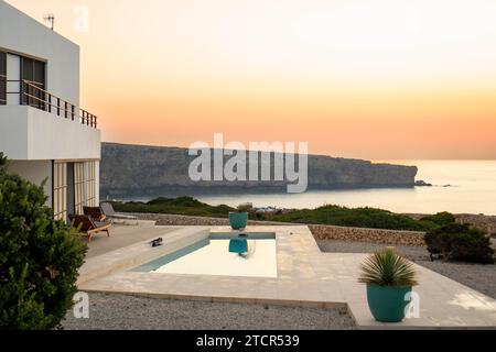 Eine luxuriöse weiße Villa mit einem Infinity-Pool, mit Blick auf das Mittelmeer bei Sonnenuntergang in Menorca, Spanien: Modernes Design, Stockfoto