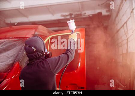 Karosseriereparatur eines Lieferwagens, die Karosserie rot lackiert ein Mechaniker in einem Atemschutzgerät einen Lieferwagen mit einer Spritzpistole Stockfoto