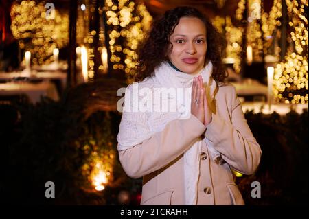 Schöne junge Erwachsene Multi-Ethno-Frau im warmen beigefarbenen Mantel, die sich Weihnachten wünscht, draußen auf dem Festplatz steht und ein glückliches fest genießt Stockfoto