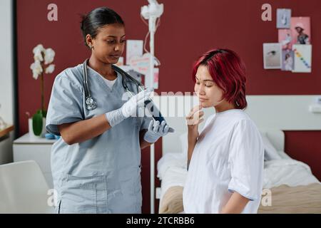 Junge professionelle plastische Chirurgin mit Spiegel vor einem Teenagermädchen mit Liftspuren im Gesicht während der Sprechstunde vor dem Eingriff Stockfoto