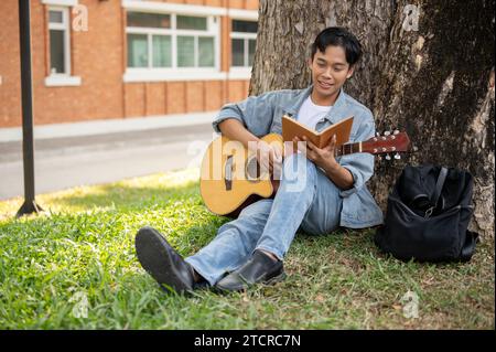 Junger positiver asiatischer Mann ruht unter dem Baum in einem Park mit seiner Akustikgitarre und übt Gitarre. Freizeit- und Hobbykonzept Stockfoto