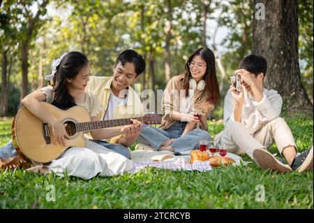 Ein hübsches junges asiatisches Paar spielt gemeinsam Akustikgitarre, während es im Sommer mit seinen Freunden ein Picknick in einem grünen Park macht. Freundschaft Co Stockfoto