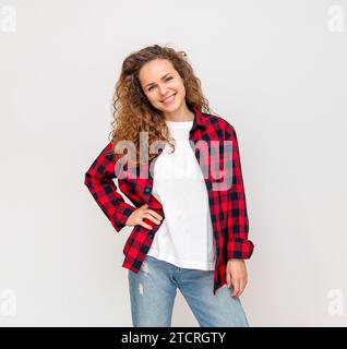 Braune Frau mit lockigem Haar, die vor einem einfachen Hintergrund steht und mit Hand auf der Taille posiert und lächelt. Stockfoto