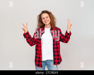 Brünette Frau mit lockigem Haar im legeren Outfit lächelt und mit Gesten versehene V-Victory-Gesten oder Friedenszeichen vor schlichtem Hintergrund. Stockfoto