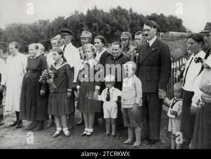 Adolf Hitler wird gezeigt, wie er während seiner Reise durch Ostpreußen eine Bauernfamilie besucht, wie der Titel andeutet. Dieser Besuch, Teil seiner Reise durch die Region, wurde wahrscheinlich inszeniert, um eine Verbindung zur ländlichen Bevölkerung zu zeigen und das Bild eines Führers zu pflegen, der sich mit allen Teilen der Gesellschaft beschäftigt. Solche Besuche waren typisch für die Propagandabemühungen der Nazis, mit dem Ziel, Hitler als eine vereinheitlichende Figur darzustellen, die soziale und wirtschaftliche Spaltungen überschritt und seine Persönlichkeit als Führer des Volkes festigte. Stockfoto