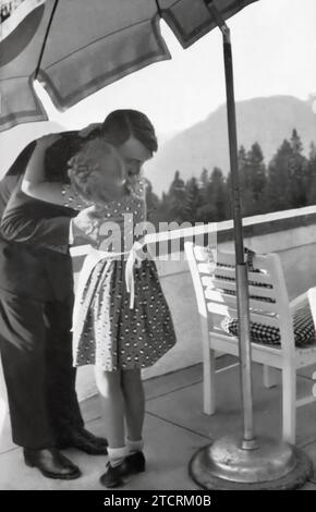 Adolf Hitler wird in einem ruhigen Moment in Obersalzberg gefangen genommen, mit einem jungen Mädchen, das ihn umarmt. Dieses Bild auf einer Terrasse mit malerischer Bergkulisse zeigt eine sanfte und mitfühlende Seite Hitlers, ein krasser Kontrast zu seinem üblichen autoritativen Verhalten. Stockfoto