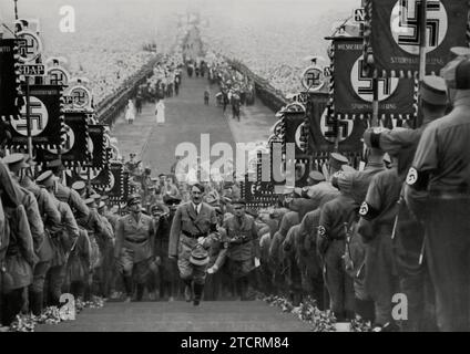 Adolf Hitler wird bei einer massiven Kundgebung der NSDAP in Bückeberg 1934 die Treppe hinaufsteigen sehen. Diese Veranstaltung, Teil des jährlichen Erntefestes, war ein bedeutendes Propagandawerkzeug, das die Fähigkeit des Regimes demonstrierte, große Menschenmengen unter seiner Ideologie zu mobilisieren und zu vereinen. Die Rallye 1934 war besonders für ihren großen Umfang und die orchestrierte Darstellung von Loyalität und Einheit innerhalb der Partei bekannt. Stockfoto