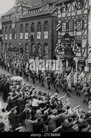 Bei der Nürnberger Kundgebung 1935, am Tag der Bundeswehr, marschieren Soldaten die Straße hinunter, die Flaggen der deutschen historischen Armee tragen, während Zuschauer und Unterstützer mit dem rechten Arm grüßen. Diese eindrucksvolle Szene unterstreicht das militärische Erbe und die Tradition, zeigt die Kontinuität und das historische Ansehen der deutschen Armee und spiegelt gleichzeitig die breite öffentliche Unterstützung und Beteiligung an der NS-Propaganda wider. Stockfoto