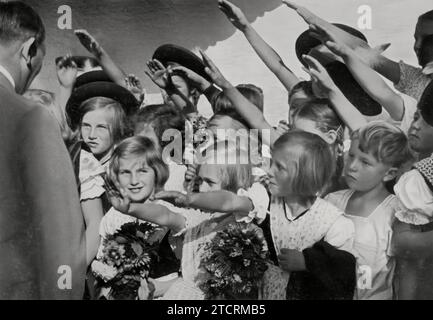 In der Kinderhände wird Adolf Hitler gesehen, wie er Kinder grüßt und Blumen von ihnen erhält. Dieses Bild ist ein ergreifendes Beispiel dafür, wie das Nazi-Regime orchestrierte Interaktionen mit Jugendlichen nutzte, um ein öffentliches Bild von Wohlwollen und Zugänglichkeit zu pflegen. Der Akt der Annahme von Blumen von Kindern war ein verbreitetes Propagandawerkzeug, das Hitlers Image weicher machen und die Idee eines pflegenden, väterlichen Führers fördern sollte, der eng mit der jüngeren Generation verbunden ist. Stockfoto
