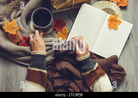 Weibliche Hände stricken Pullover auf dem Boden mit Herbstblättern und einer Tasse Kaffee Stockfoto