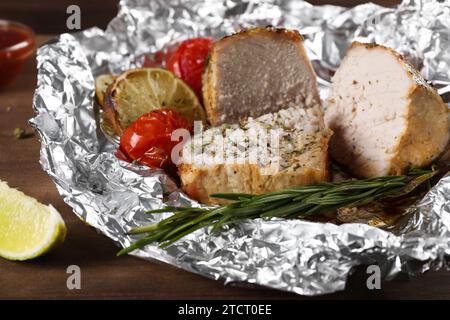 Fleischstücke in Folie gebacken mit Tomaten auf Holztisch, Nahaufnahme Stockfoto