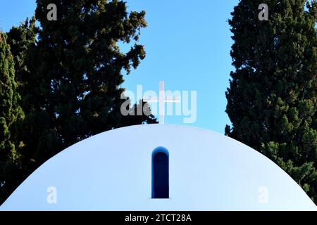 Nahaufnahme der alten weißen griechischen Kapelle, kleine Kirche mit glänzendem weißem Kreuz in natürlichem Sonnenlicht, blauem Himmel und grünen Zypressen im Hintergrund. Stockfoto