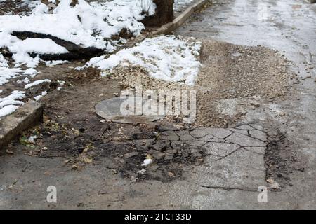 Unerledigte Straßenreparaturen. Kanalluke und gebrochener Asphalt, bedeckt mit zerquetschtem Stein. Schmutz und Schnee auf einer kaputten Straße in einem Wohngebiet. Flach Stockfoto