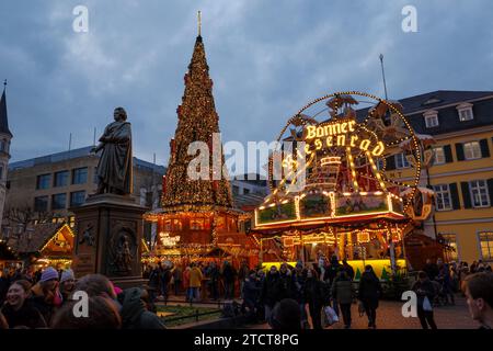 Bonn, Deutschland - 6. Dezember 2023: Blick in die Dämmerung auf einen belebten Weihnachtsmarkt mit festlichen Lichtern, einem Karussell und einem großen geschmückten Weihnachtsbaum dahinter Stockfoto