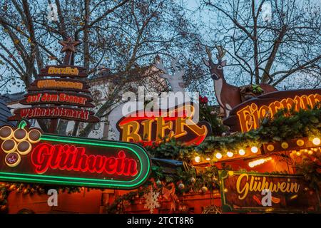 Bonn, Deutschland - 6. Dezember 2023: Dämmerungsblick auf einen festlichen Weihnachtsmarkt mit beleuchteten Schildern für Bier und Glühwein. Stockfoto