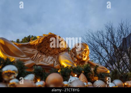 Bonn, Deutschland - 6. Dezember 2023: Goldene Engelsfigur inmitten festlicher Dekorationen mit einem verschwommenen Hintergrund von Bäumen und Dämmerungshimmel. Stockfoto