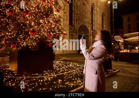 Seitenporträt einer touristischen Frau, die Como in Italien besucht, mit Smartphone, Foto von einem schönen Weihnachtsbaum mit festlichen Lichtern A Stockfoto