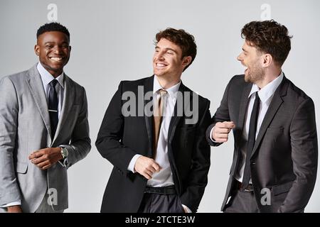 Gut aussehende, fröhliche verschiedene männliche Models in eleganten Anzügen, die aufrichtig vor grauem Hintergrund lächeln Stockfoto