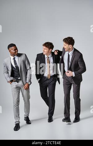 Gut aussehende glückliche junge männliche Models in schicken Anzügen, die aufrichtig vor grauem Hintergrund lächeln Stockfoto