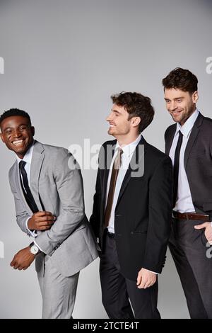 Gut aussehende, fröhliche multikulturelle männliche Models in eleganten Anzügen, die aufrichtig vor grauem Hintergrund lächeln Stockfoto