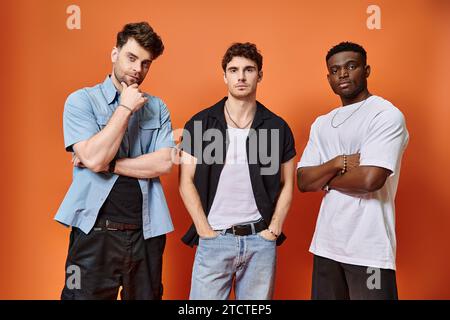 Drei gut aussehende multikulturelle Freunde in legeren urbanen Outfits posieren zusammen vor orangefarbenem Hintergrund Stockfoto
