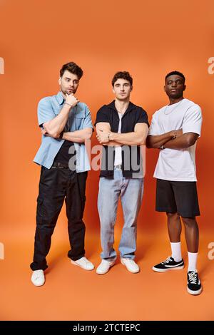 Drei gutaussehende multirassische Freunde in lässigen urbanen Outfits posieren zusammen vor orangefarbenem Hintergrund Stockfoto