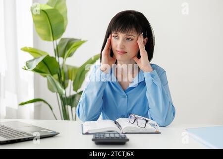 Überarbeitete, müde junge Geschäftsfrau massiert Schläfen, leidet an Kopfschmerzen nach der Computerarbeit sitzt am Schreibtisch mit Laptop im Büro, erschöpfte Frau mit geschlossenen Augen, die den Kopf berührt, lindert Schmerzen Stockfoto