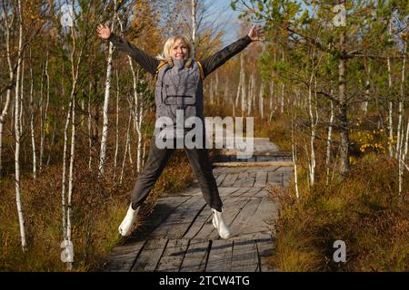 Eine glückliche Frau mit Rucksack springt auf einem Holzweg in einem Sumpf in Jelnya, Weißrussland. Stockfoto