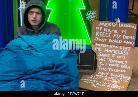 Windsor, Großbritannien. Dezember 2023. Reece, ein Mann, der nach dem Tod seines Vaters obdachlos geworden ist, sitzt auf dem kalten Boden im Regen vor einem Geschäft im wohlhabenden Windsor, Berkshire, in der Hoffnung, dass Passanten ihm ein paar Münzen geben. Polly Neate, Chief Executive von Shelter, sagte Sky News, dass Obdachlosigkeit auf der Weihnachtsliste von niemandem steht, aber 309.000 Menschen werden diese Zeit des Jahres in einem winzigen Hostel-Zimmer verbringen oder in einer Tür frieren. Quelle: Maureen McLean/Alamy Live News Stockfoto