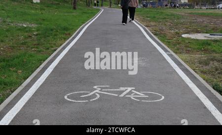 Asphaltradweg in einem Park mit sichtbaren Beinen von zwei Fußgängern, ein Fahrradschild auf der Straßenoberfläche in einem öffentlichen städtischen Raum Stockfoto