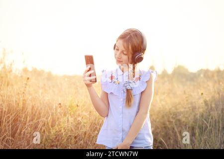 Grillendes Teenager-Mädchen mit Kopfhörern, die das Smartphone in der Hand halten Auf Sunshine Field Hintergrund Stockfoto