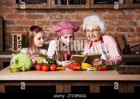 Oma mit zwei Enkelinnen, die Rezepte aus dem Kochbuch lesen und Salat kochen Stockfoto