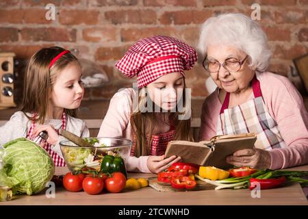 Oma mit zwei Enkelinnen, die Rezepte aus dem Kochbuch lesen und Salat kochen Stockfoto