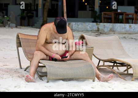 Mann im Kofferraum mit fettem Bauch am Strand mit Smartphone in den Händen. Übergewicht und All-Inclusive-Konzept Stockfoto