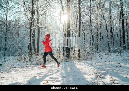 Trailläuferin in leuchtend roter, winddichter Jacke, die an einem sonnigen, frostigen Tag einen malerischen, verschneiten Waldweg joggen lässt. Sportlich aktive Menschen und Winter Stockfoto