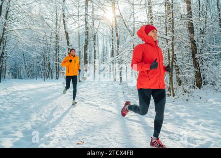 Junge Paare Trailläufer Mann und Frau kleideten helle winddichte Jacken, die während des sonnigen frostigen Tages im malerischen verschneiten Wald laufen. Sportlich aktiv p Stockfoto