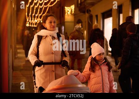 Junge Mutter schiebt Kinderwagen, geht mit ihrer Tochter auf der Stadtstraße, genießt die festliche Atmosphäre des Weihnachtsfestplatzes im Nig Stockfoto